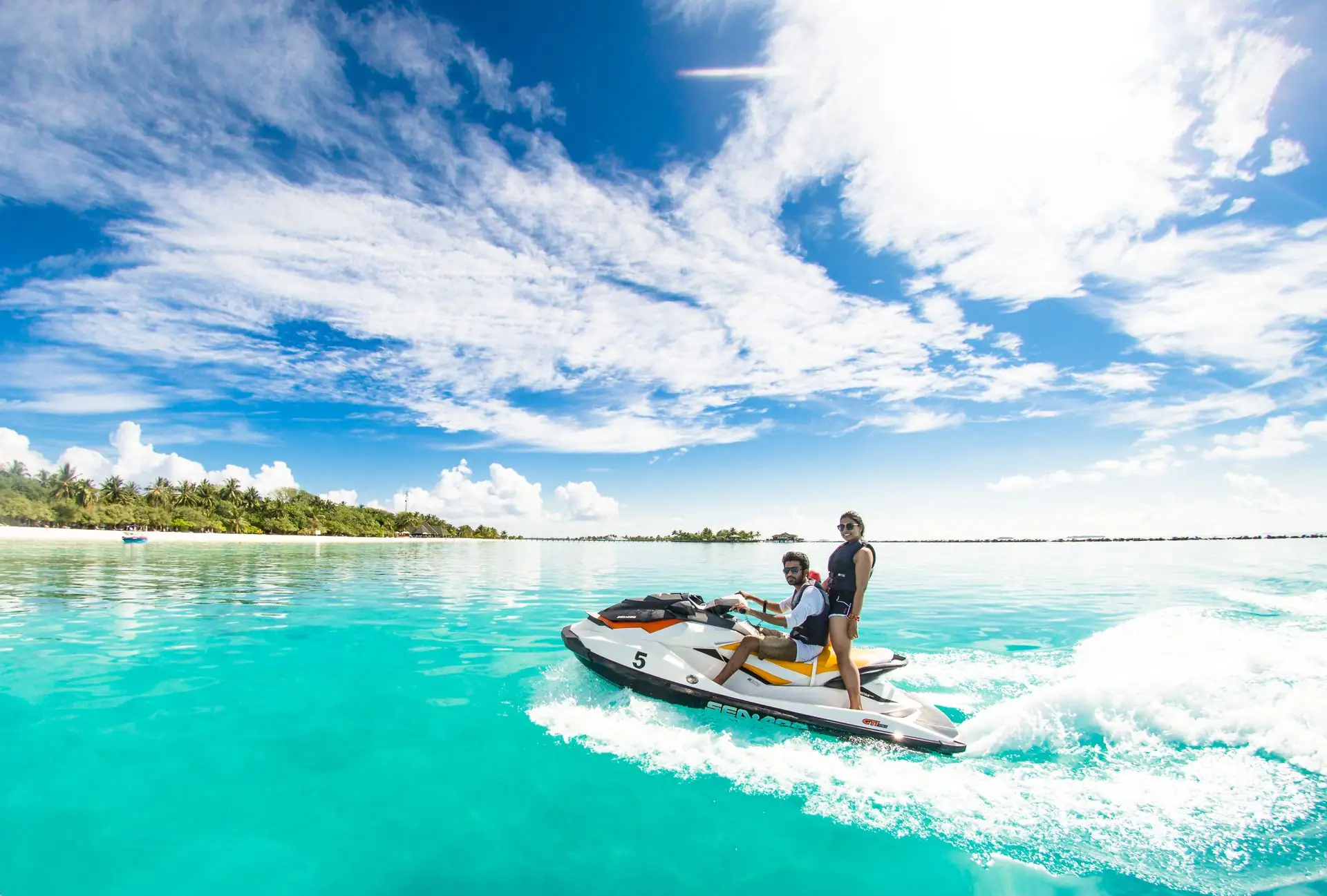 A couple speeding on a jet ski across turquoise water by a palm-fringed beach